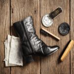 A black leather boot sits on a wooden surface, surrounded by an array of shoe polishing tools. Among them are two types of shoe brushes essential for maintaining shine, alongside a soft cloth and tins of white and black polish.