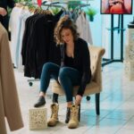 A woman sits on a chair in a clothing store, trying on tan boots. Surrounded by racks of clothes and shoes, she ponders shoe care tips for extending shoe life. The store boasts a tiled floor and modern, stylish interior design.