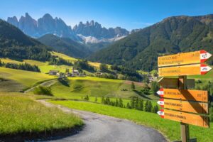 A winding path leads through lush green meadows towards scattered houses in a valley, set against jagged mountain peaks under a clear blue sky. Wooden signposts mark hiking trails with colorful directional arrows and trail names in German, perfect for those planning an Italian road trip.